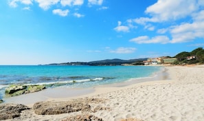 A beach with clear blue water, sandy shore, and buildings in the background. - Villa with private pool (Alghero)