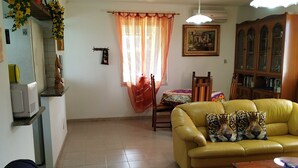 A living room with a yellow sofa, a blue couch, a wooden coffee table, and a red rug. There is a window with orange curtains and a painting on the wall.