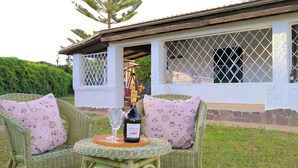 A wicker chair with a bottle and glass on a table, a white building with a window, and a hedge in the background.