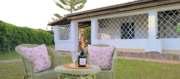 A wicker chair with a bottle and glass on a table, a white building with a window, and a hedge in the background. - Villa with private pool (Alghero)