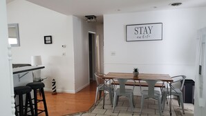 Un espace repas avec une table en bois et des chaises en métal, une petite plante en pot et un panneau fixé au mur sur lequel on peut lire : « ATTENDEZ ici pendant que vous patientez ».