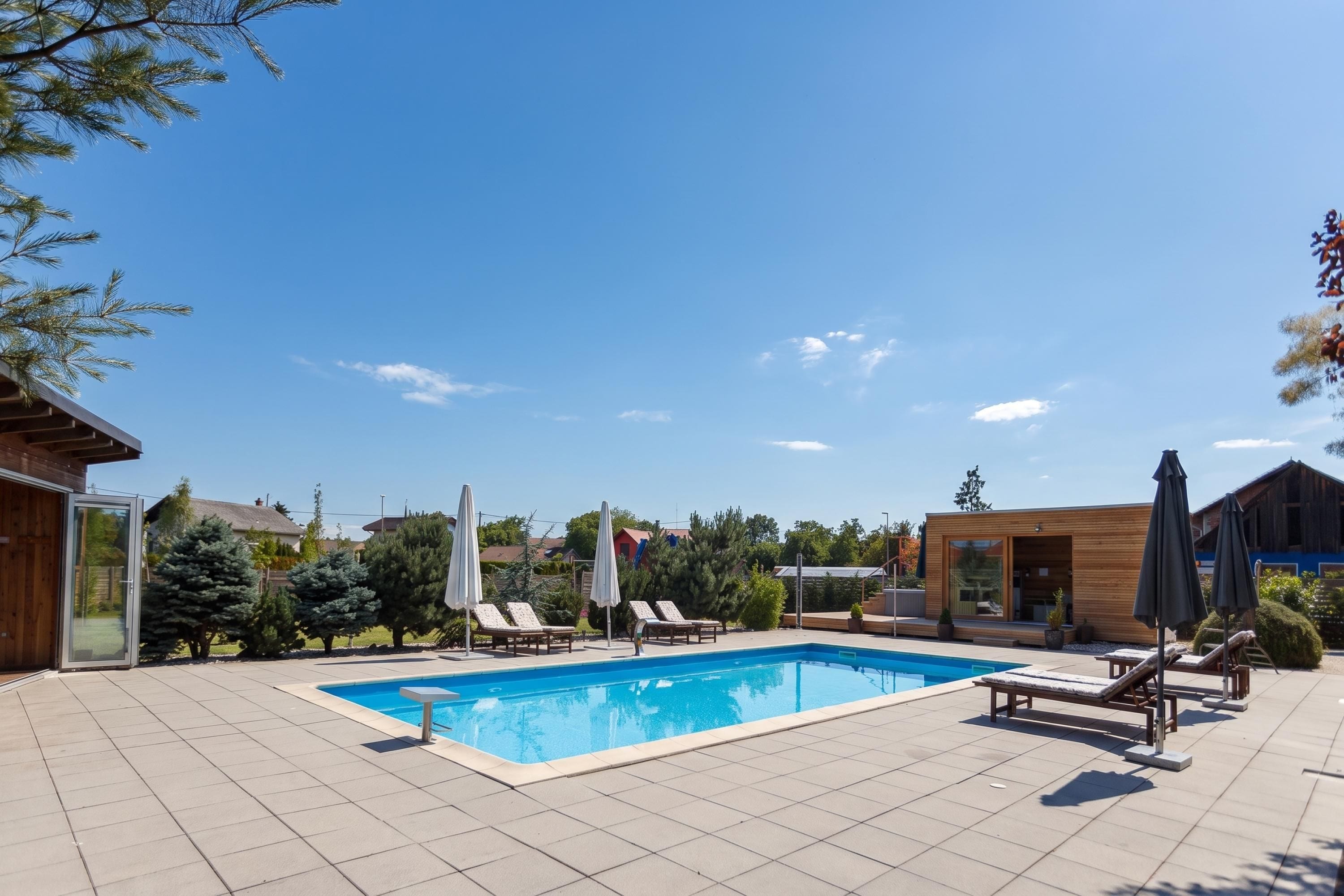 Piscina con sillas de descanso y sombrillas, rodeada de árboles y edificios.
