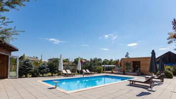 Piscina con sillas de descanso y sombrillas, rodeada de árboles y edificios.