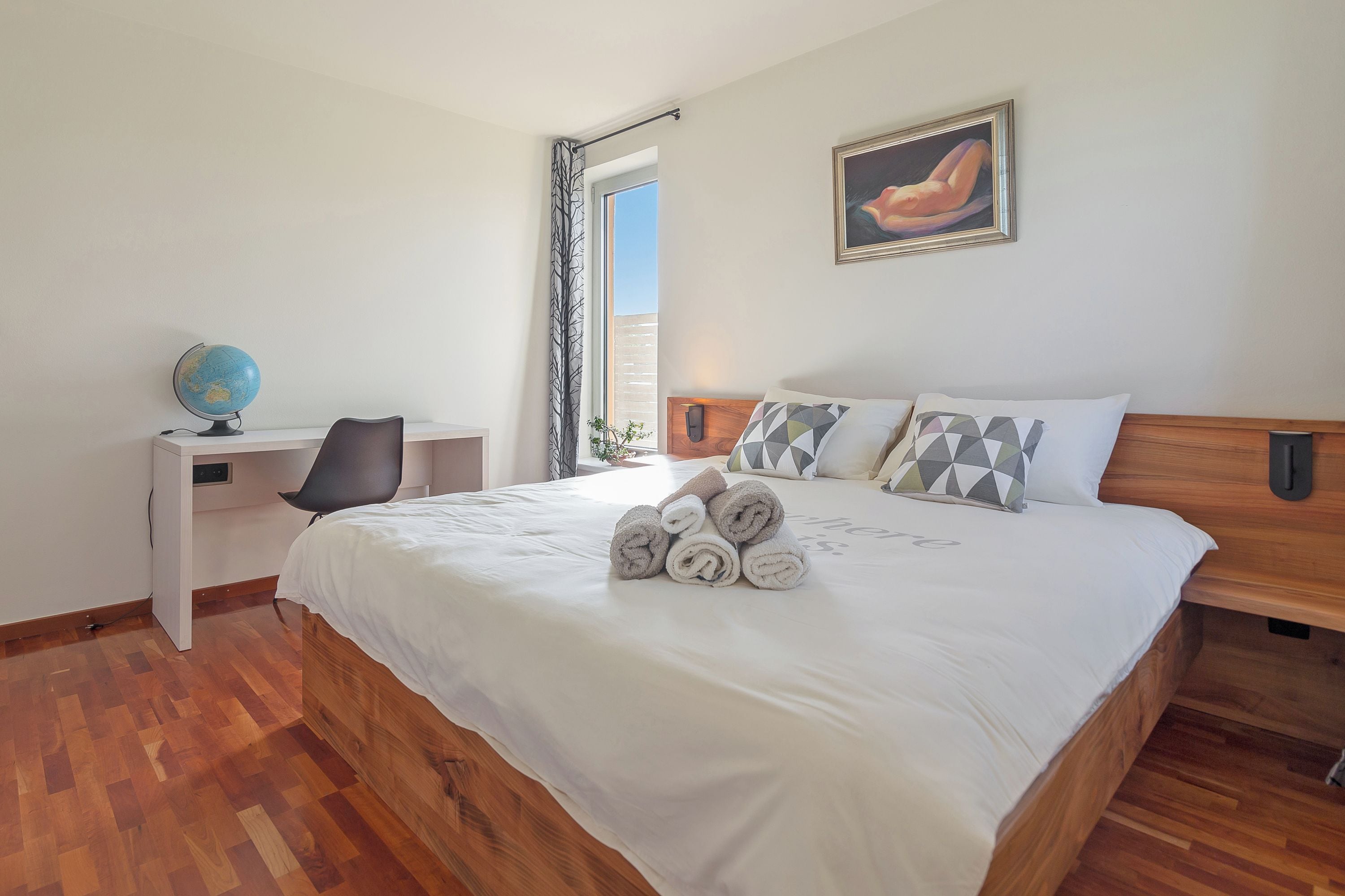 Una cama bien hecha con toallas enrolladas, un globo terráqueo, una silla y un cuadro en la pared.