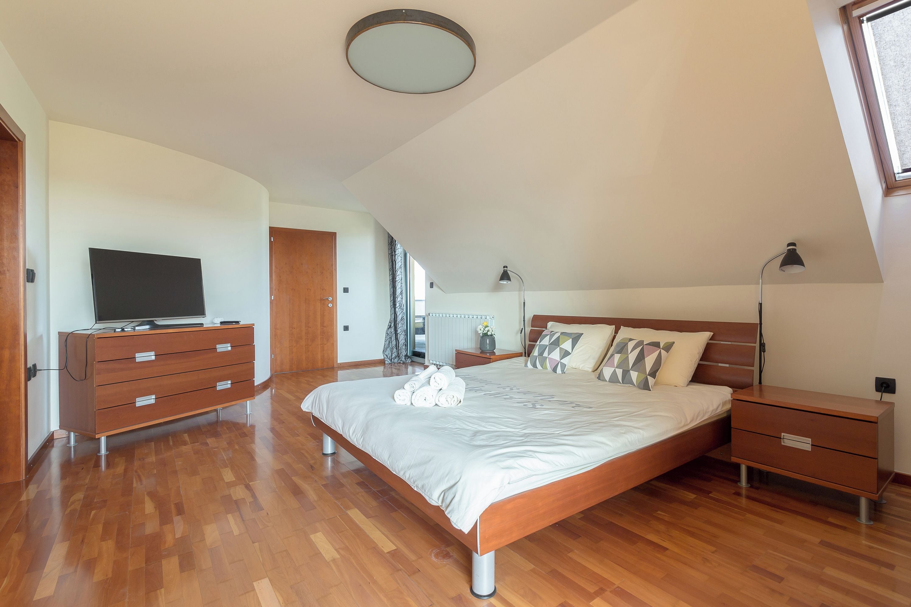 Un dormitorio con una cama, un tocador, una mesita de noche, una lámpara y un televisor.
