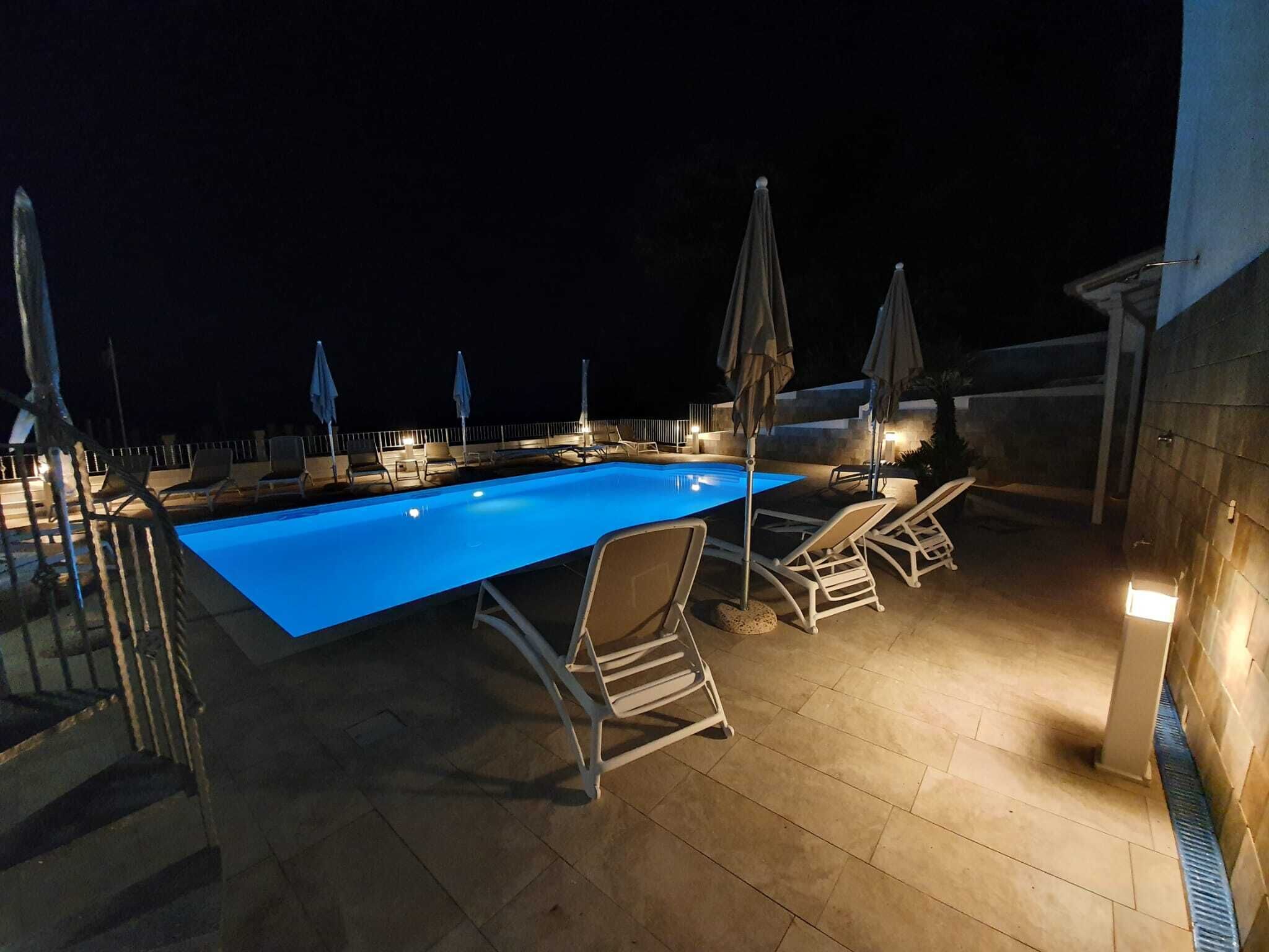 Una piscina al aire libre con sillas de descanso y sombrillas, rodeada por un muro de piedra y una barandilla de metal.