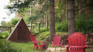 En træhytte med et trekantet tag, omgivet af skov. Tre røde Adirondack-stole og en stenvæg ligger i nærheden.
