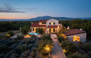 Une maison en pierre à deux étages, dotée d’une piscine, entourée d’oliviers et surplombant un paysage montagneux.