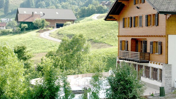 A two-story wooden house with a balcony, surrounded by lush greenery and a winding road. - A large chalet for getting together with family, friends or colleagues!  (Cerniat)