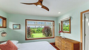A bedroom with a bed, a dresser, a ceiling fan, a window with a view of greenery, and a framed picture on the wall. - Oceanfront Hanalei Bay home - Hanalei Surf (Hanalei)