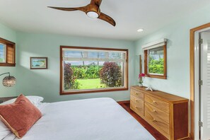 Ein Schlafzimmer mit einem Bett, einer Kommode, einem Deckenventilator, einem Fenster mit Blick auf Grünflächen und einem gerahmten Bild an der Wand.