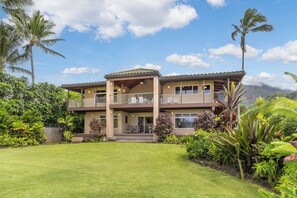 Une maison à deux étages avec un balcon, entourée d’une végétation luxuriante et sous un ciel dégagé.