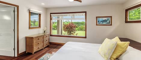 Un dormitorio con una cama grande, un ventilador de techo, una ventana con vistas a zonas verdes y un aparador de madera.