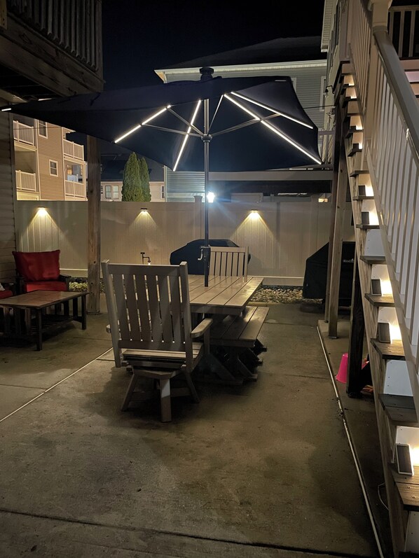Un patio al aire libre con mesa y sillas de madera, una parrilla y una zona de estar cubierta con luces.