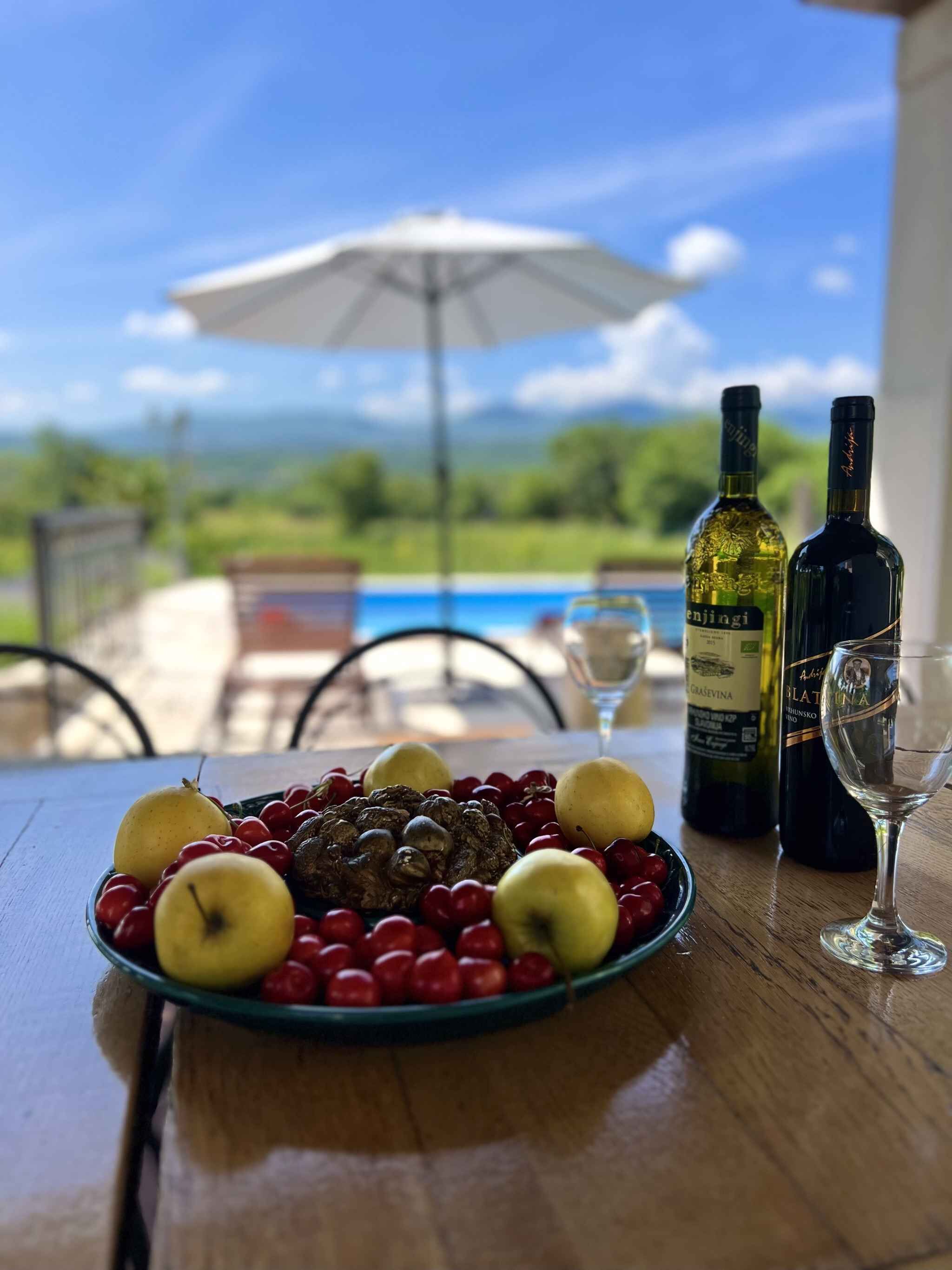 Et bord med frugt, to vinflasker og et glas, med udsigt over et smukt udendørs landskab og en hvid parasol.