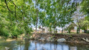 A serene riverside view with a wooden bridge, stone steps, and a row of cabins with a stone wall in the background. - Boutique Tiny Home - River, Fire Pit, Walkable (Waynesville)