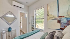 A bedroom with a bed, a dresser, a mirror, an air conditioner, a window with blinds, and a painting on the wall. - Boutique Tiny Home - River, Fire Pit, Walkable (Waynesville)