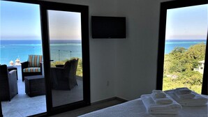 A room with a bed, a TV, and a balcony with wicker furniture and a view of the sea.