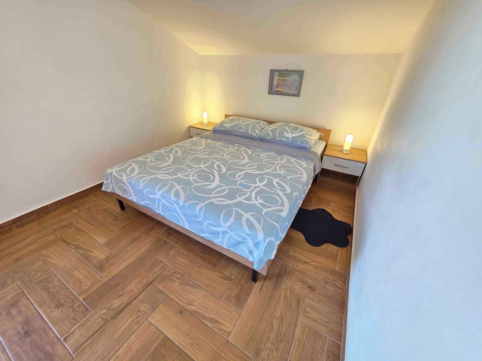A bedroom with a herringbone-patterned wooden floor, a bed with a white and gray patterned bedspread, two bedside tables with lamps, and a framed picture on the wall.
