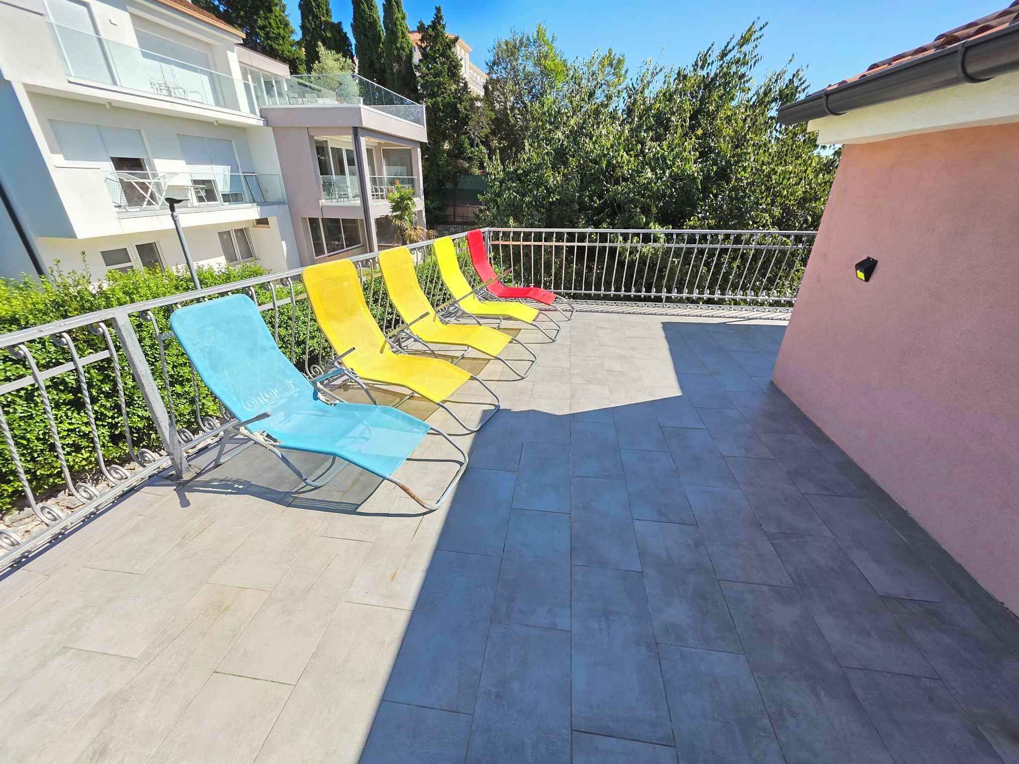 Terraza en la azotea con varios sillones de sol de diferentes colores, barandilla de metal y un edificio al fondo.