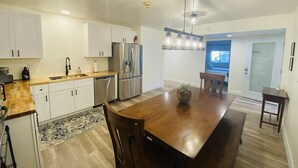 A modern kitchen with white cabinets, a stainless steel refrigerator, a wooden dining table, and pendant lights. - Historic Firehouse - Spacious, Private & Clean (Kachina Village)