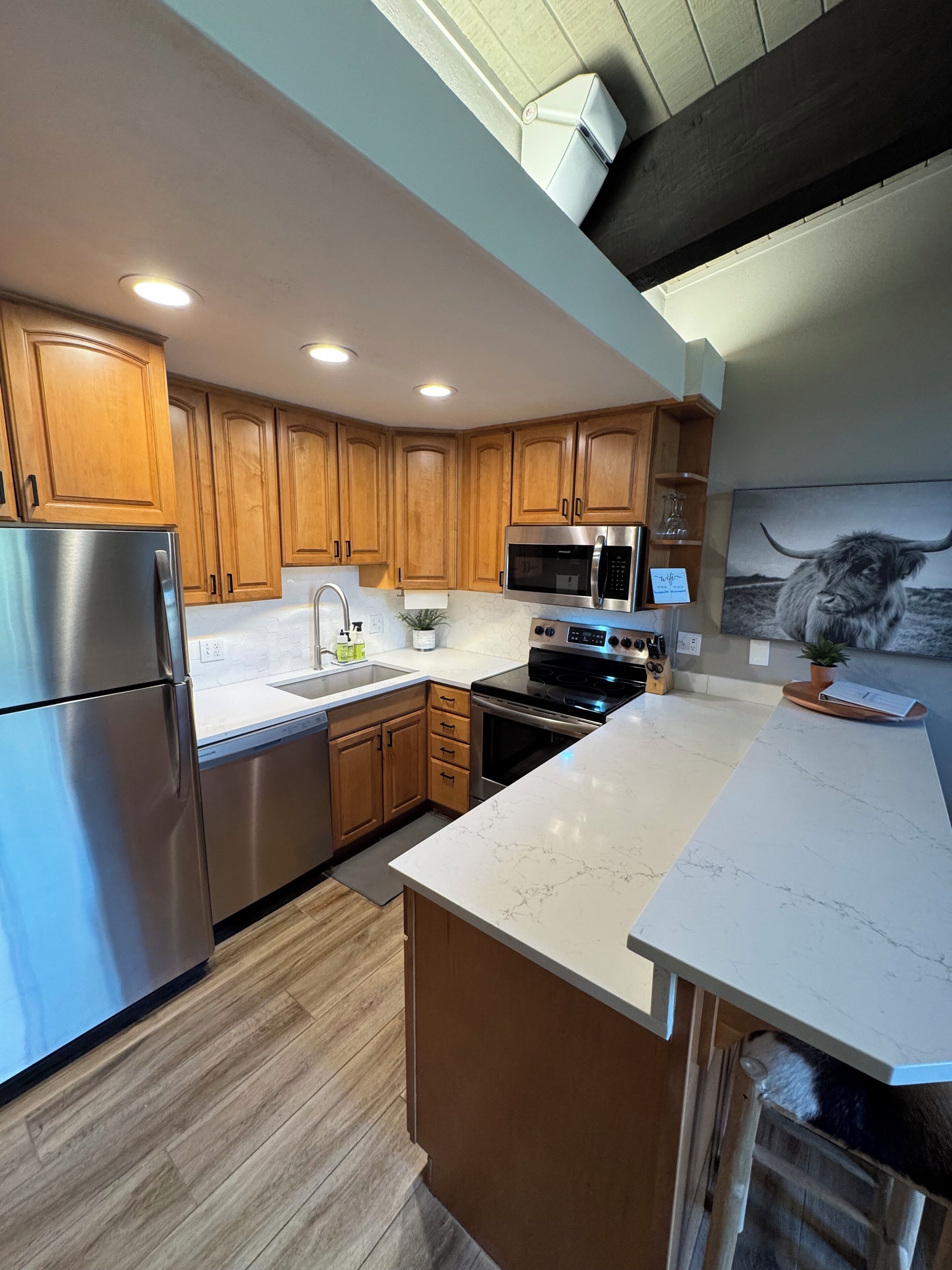 A modern kitchen with wooden cabinets, stainless steel appliances, a marble countertop, and a black and white artwork on the wall.