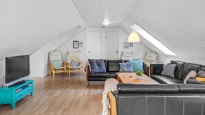 Tilava ullakkoasunto, jossa on korkea katto, puiset kattopalkit, musta nahkasohva, puinen sohvapöytä ja televisio turkoosinvärisellä jalustalla.