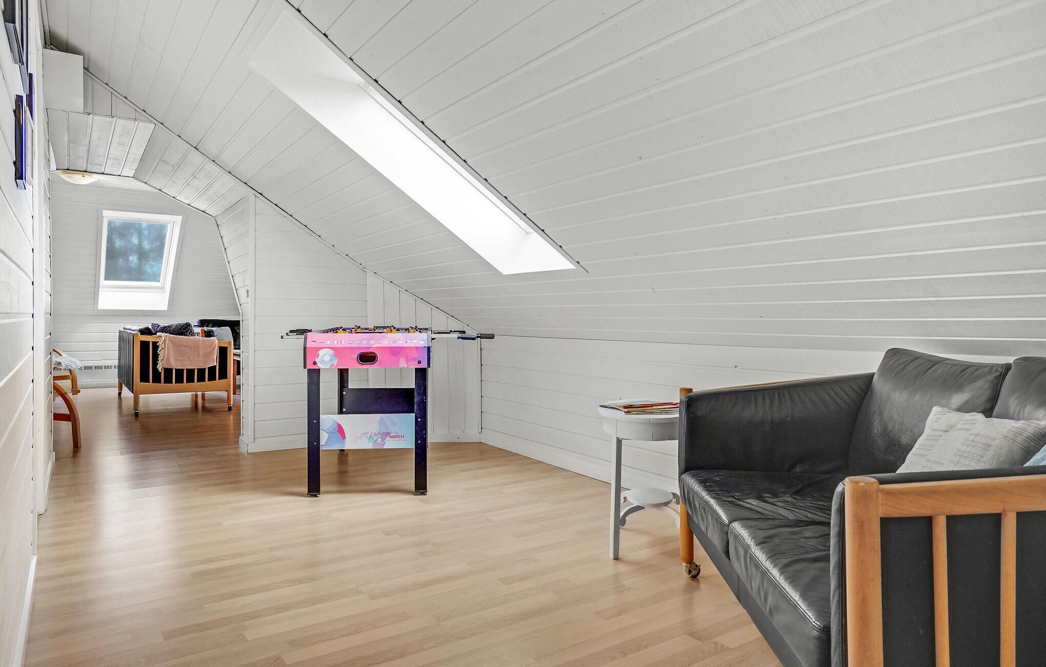 A spacious attic room with a high ceiling, wooden flooring, a black leather sofa, a small table with a pink and blue machine, and a skylight.