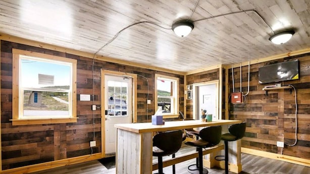 Una habitación rústica de estilo cabaña con paredes de madera, una mesa de madera y dos sillas. Cuenta con ventanas amplias, una puerta y un televisor montado.