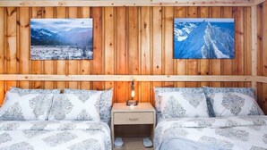 Una habitación con paredes de paneles de madera, dos camas con ropa de cama estampada, una mesita de noche y dos cuadros con paisajes de montañas nevadas.