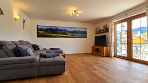 Un salon avec un canapé d’angle gris, un téléviseur à écran plat posé sur un meuble en bois, un grand tableau représentant un paysage et une porte-fenêtre coulissante donnant sur un espace extérieur.