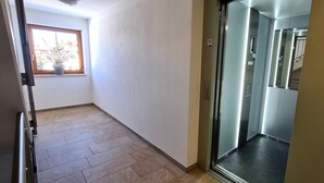 Un couloir doté d’un sol carrelé, une fenêtre avec un pot de fleurs et un ascenseur avec une porte en verre.