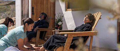 A spa setting with wooden chairs, a massage table, and a small table with a bowl of fruit and a vase of flowers.