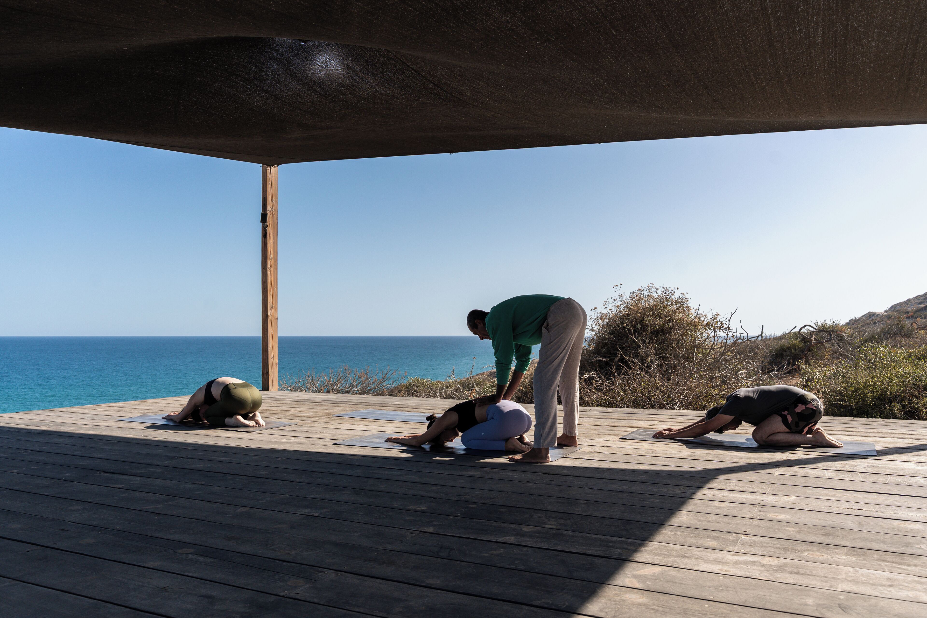 A wooden deck with yoga mats, a person assisting another, and a view of the ocean.