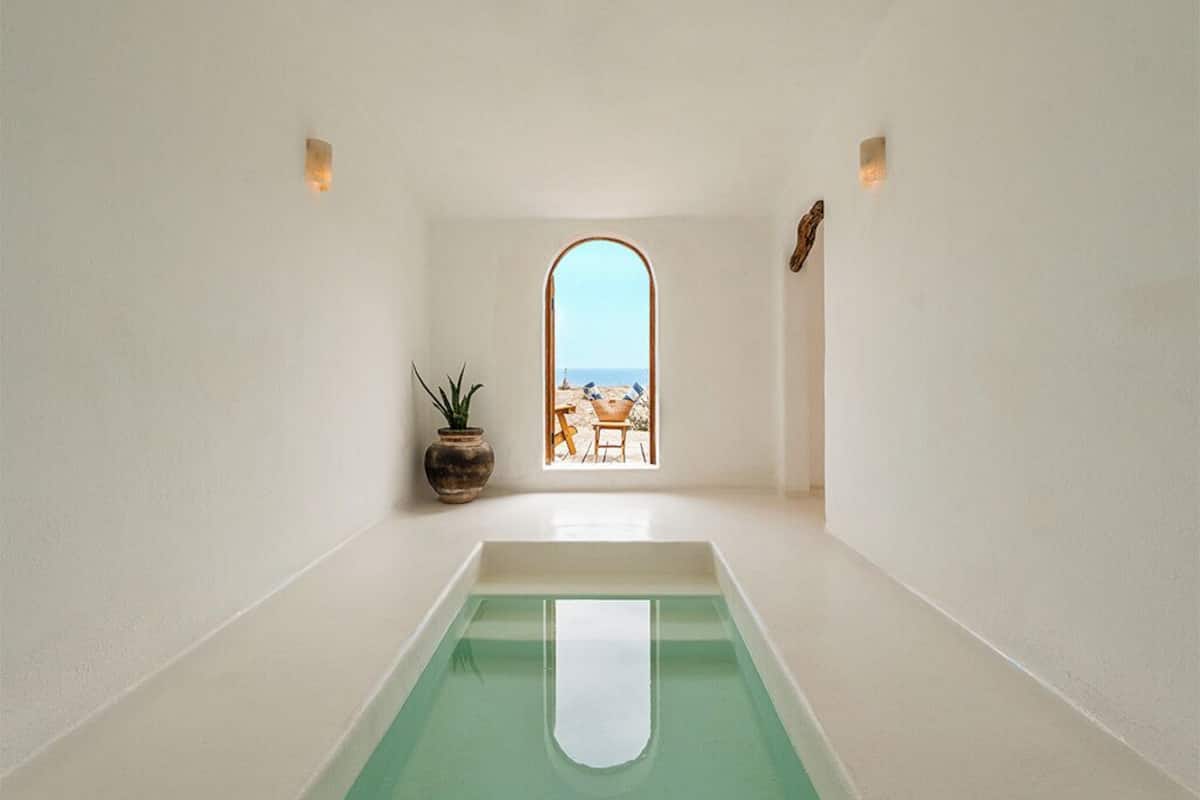 A minimalist room with a central pool, a potted plant, and a view of the beach through a window.