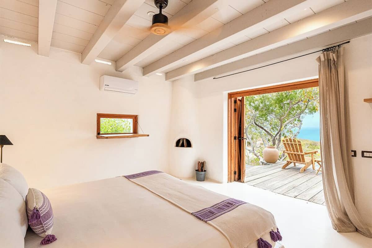 A bedroom with a large bed, a window, an air conditioning unit, and a view of the outdoors.