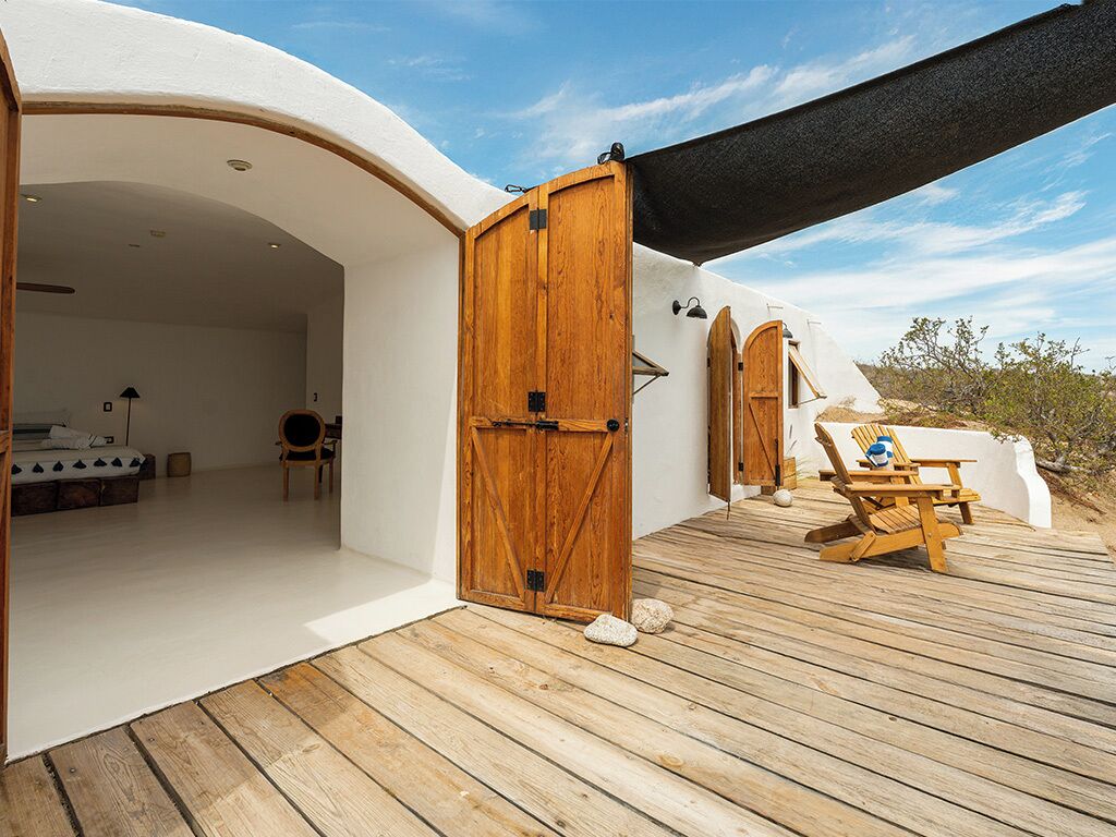 Une terrasse en bois avec une porte en bois donnant sur une pièce où se trouvent un lit et une chaise.