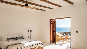 A bedroom with a bed, a wooden headboard, a nightstand, a lamp, and a view of the sea through an open door. - NEST Baja, Member of Design Hotels (La Fortuna)