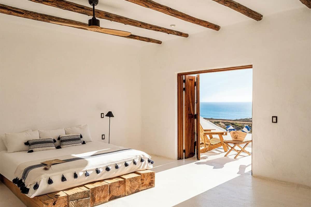A bedroom with a bed, a wooden headboard, a nightstand, a lamp, and a view of the sea through an open door.