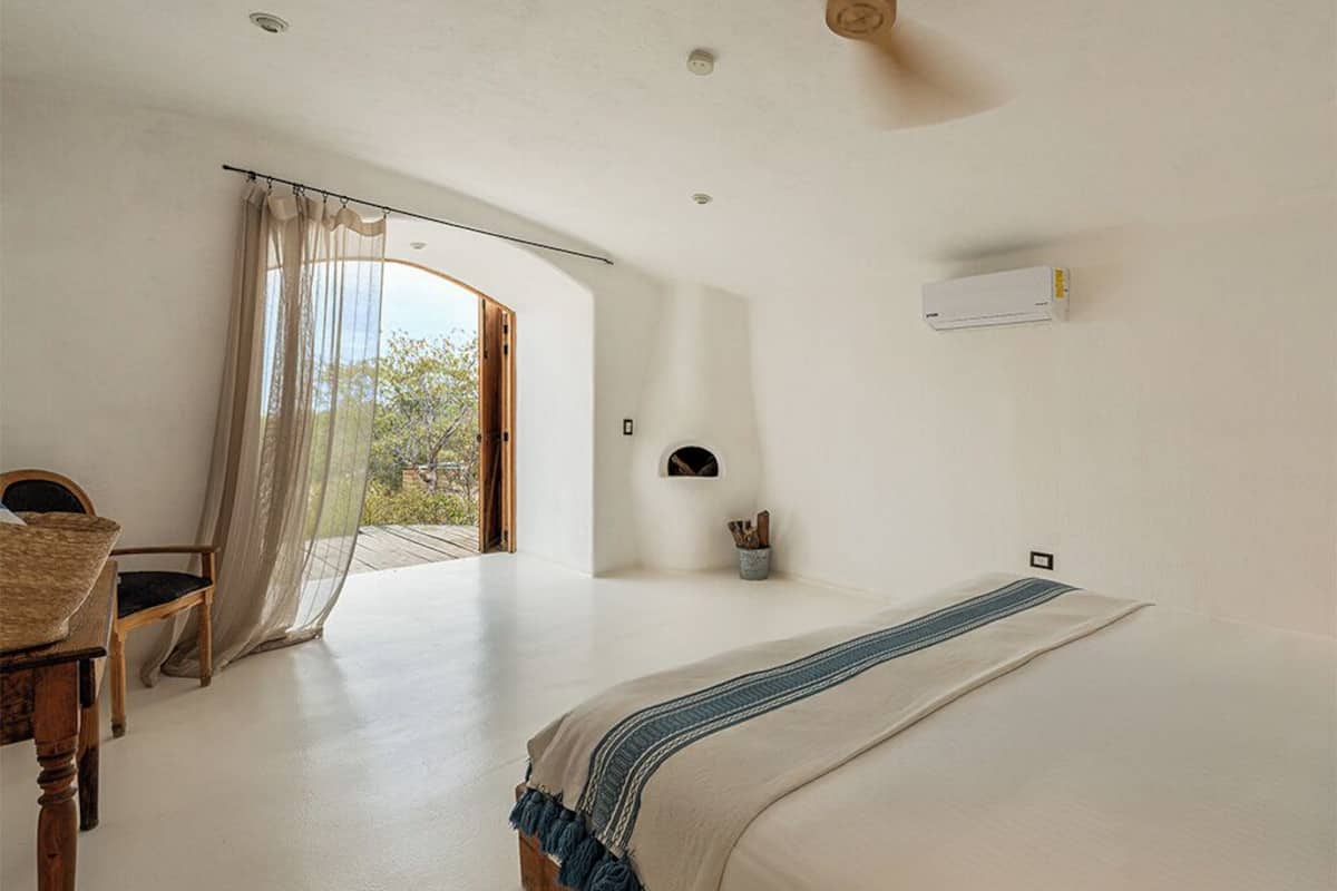 A bedroom with a bed, a chair, a window with sheer curtains, and a ceiling fan.
