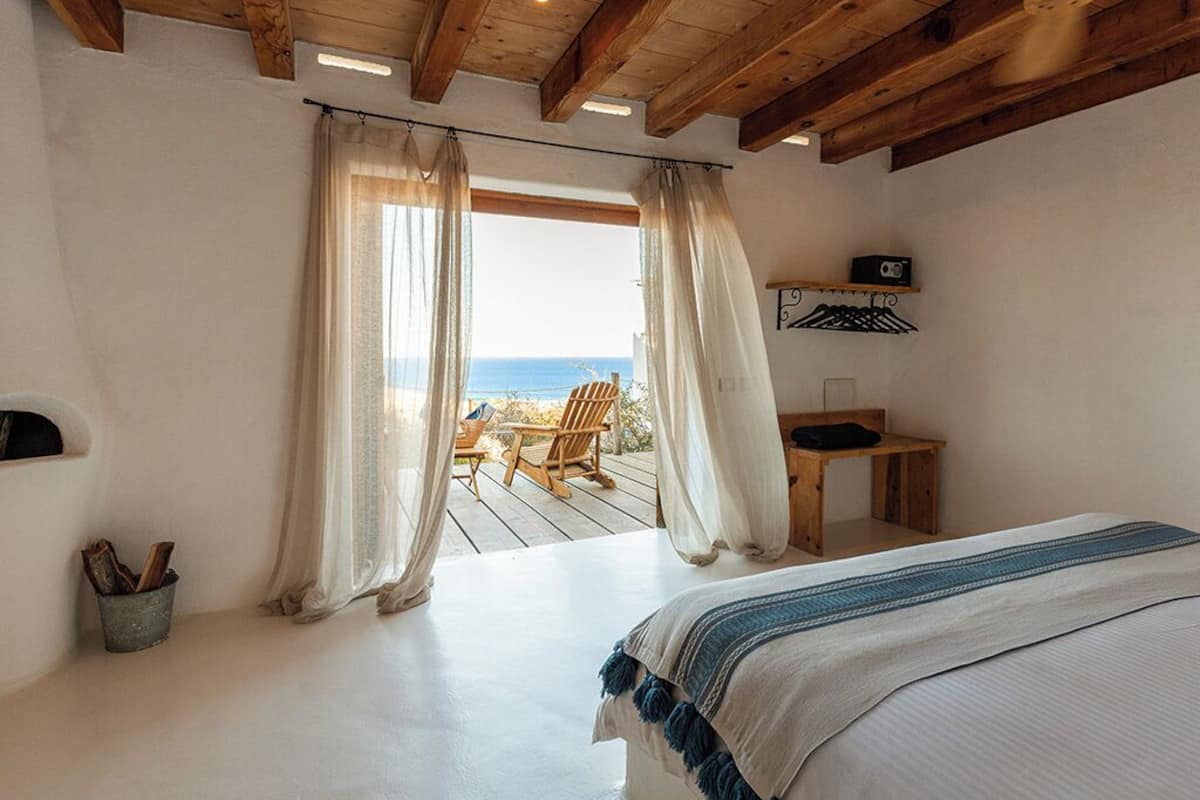 A bedroom with a large bed, wooden ceiling beams, a view of the beach, and a small table with a chair outside.