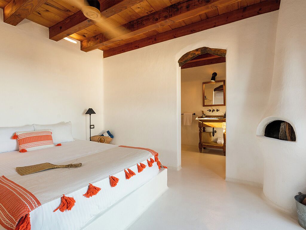 Une chambre à coucher avec un grand lit, des poutres apparentes en bois au plafond, une petite table de chevet, une lampe et une cheminée rustique.