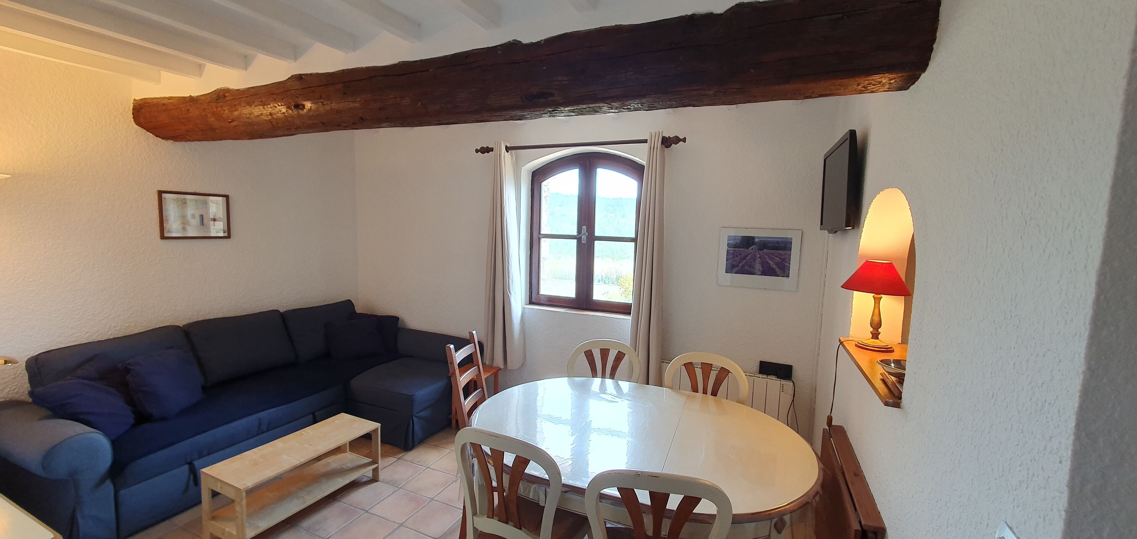 Un accogliente soggiorno con un divano blu scuro, una panca in legno, un tavolo da pranzo rotondo con sedie bianche e una finestra con tende.