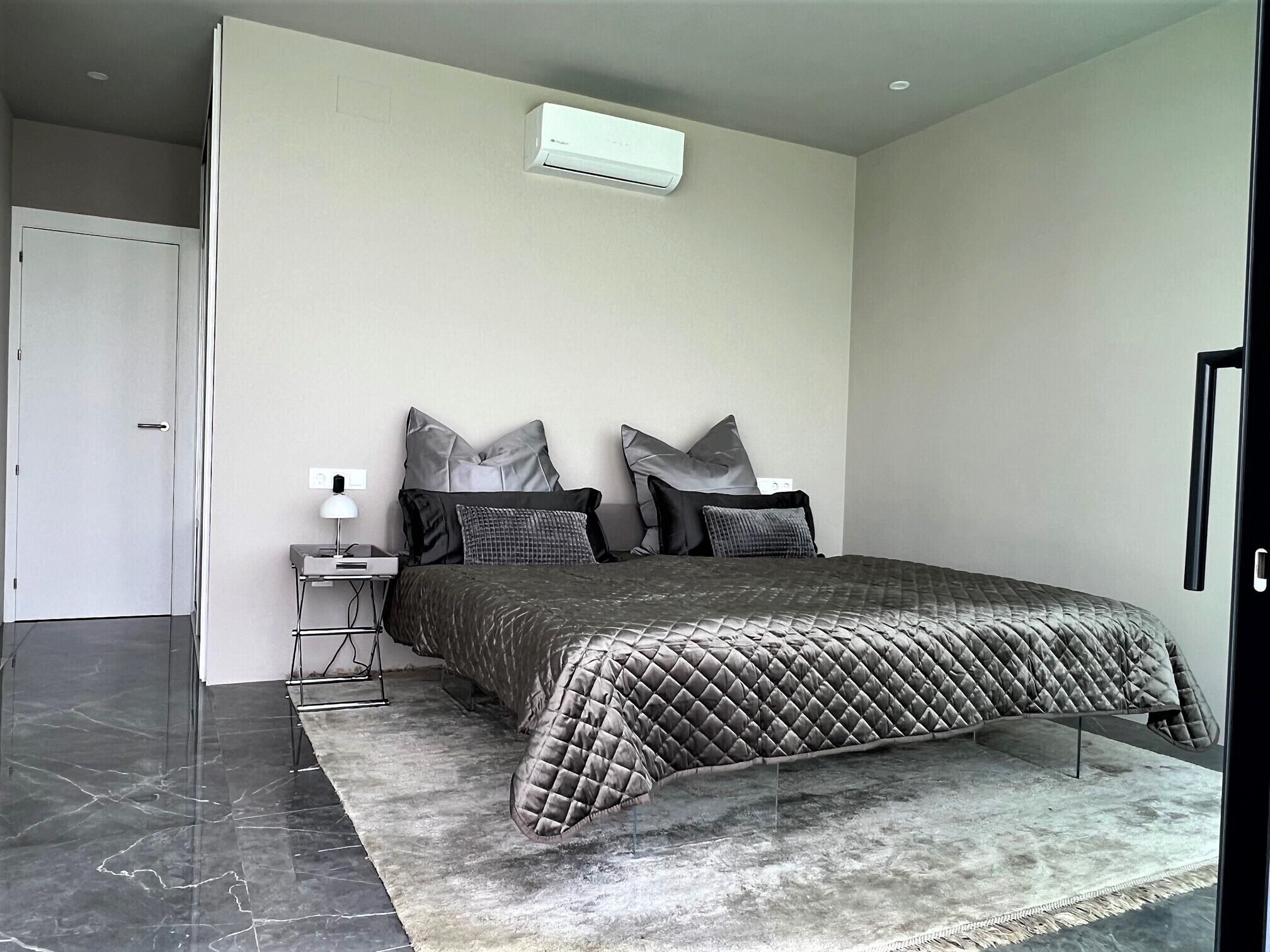 A modern bedroom with a bed, a bedside table, a lamp, and a wall-mounted air conditioner.