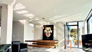 A modern living room with a large window, a colorful abstract painting, a wooden bench, and a yellow ottoman.