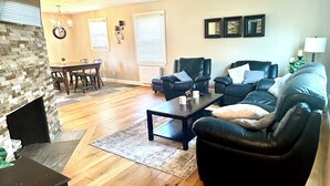A living room with a fireplace, a dining area, a black leather sofa, a coffee table, and a ceiling fan. - Beautiful Renovated Home Walk to University of Maine in Orono (Orono)