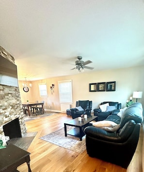 A living room with a fireplace, a dining area, a black leather sofa, a coffee table, and a ceiling fan.