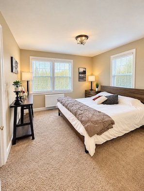 A bedroom with a bed, bedside tables, a radiator, and two windows with blinds.