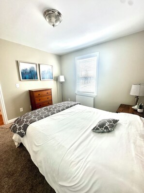 A bedroom with a bed, a dresser, a lamp, a window with blinds, and two wall-mounted pictures.