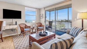 A spacious living room with a large window, a flat-screen TV, wicker chairs, a sofa, and a balcony with a view of the water. - Beach & Tennis 1604 (Bonita Springs)